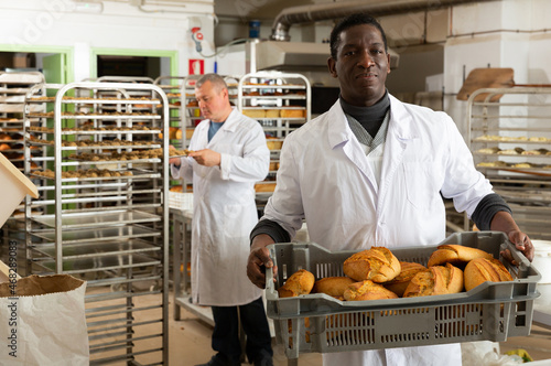 Confident African American bakery worker carrying plastic box with freshly baked loaves..