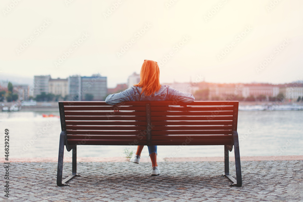 Redhead woman relaxing on bench at riverside in sunlight - obrazy, fototapety, plakaty 