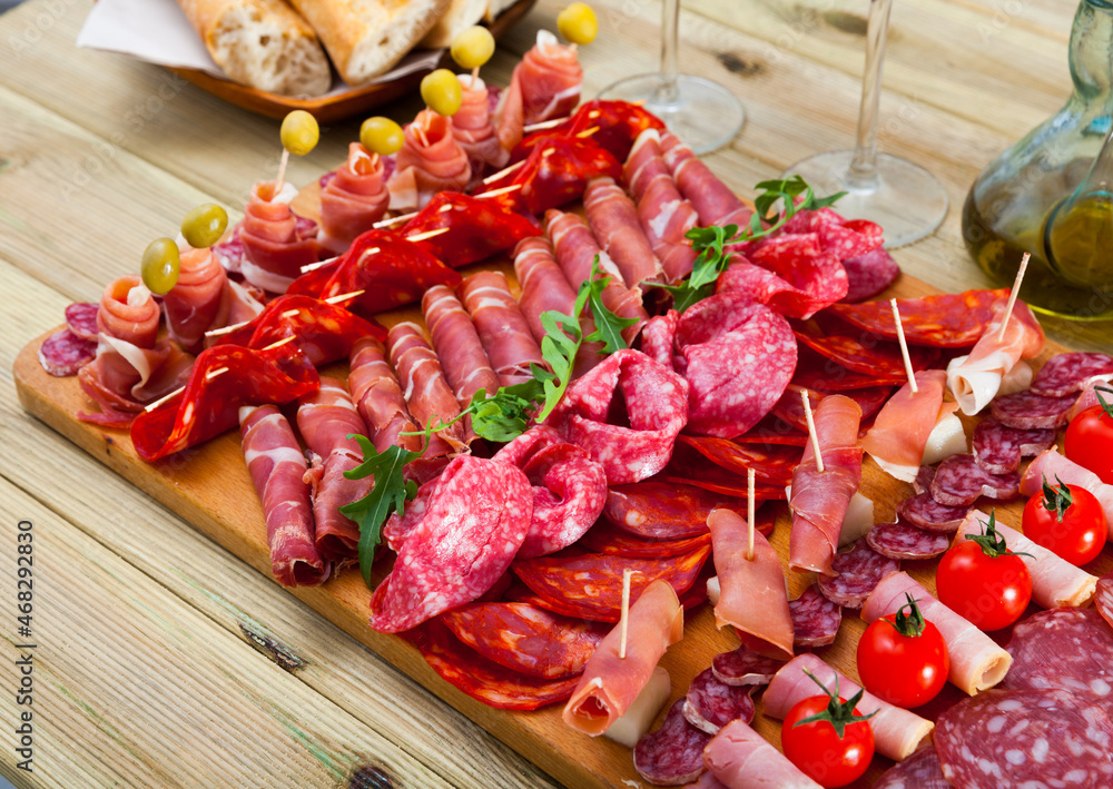 Slices of Spanish dry-cured gammon, variety of sausages and bacon on wooden board garnished with vegetables