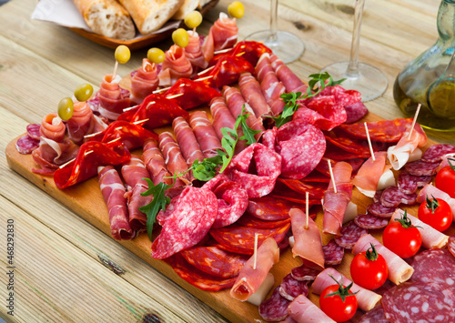 Slices of Spanish dry-cured gammon, variety of sausages and bacon on wooden board garnished with vegetables