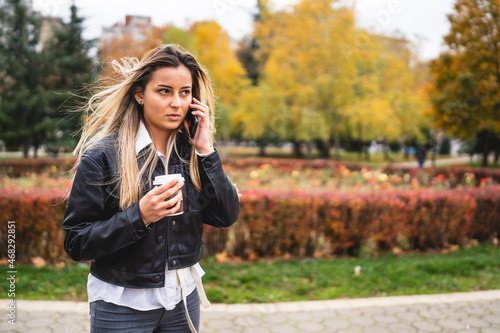 One Young caucasian woman standing in park outdoor in autumn or winter day using mobile phone to talk making a phone call receiving bad news serious holding a cup of coffee copy space real people