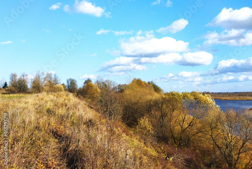 Beautiful autumn landscape in clear, sunny weather. Yellowed grass and trees on the steep bank of a blue river under a bright, blue sky with clouds. 