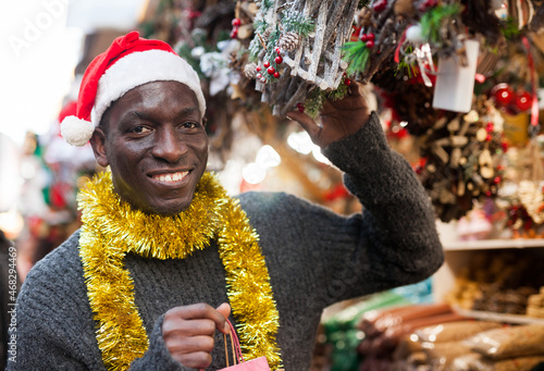 African-American guy wearing warm sweater and Santa hat walking through street Christmas fair in festive mood, choosing handmade decorations