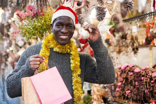 Portrait of smiling African American man in Santa hat with shopping bags on festive fair before Christmas.