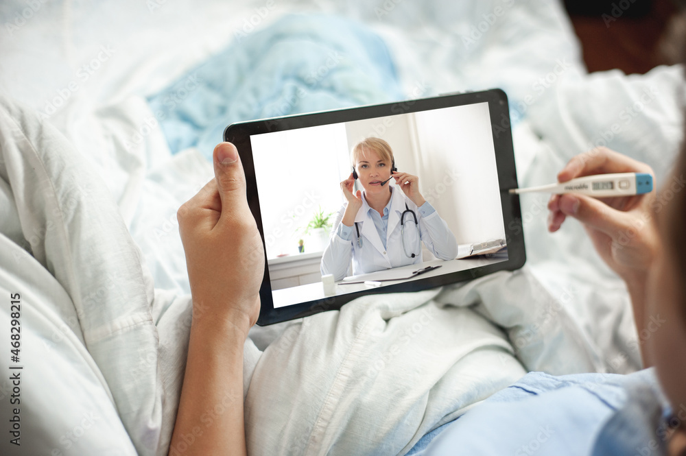 Telemedicine and online consultation concept. A young man or teenager sits in bed with an electronic thermometer and a tablet in his hands. The young man has video chat with a doctor.