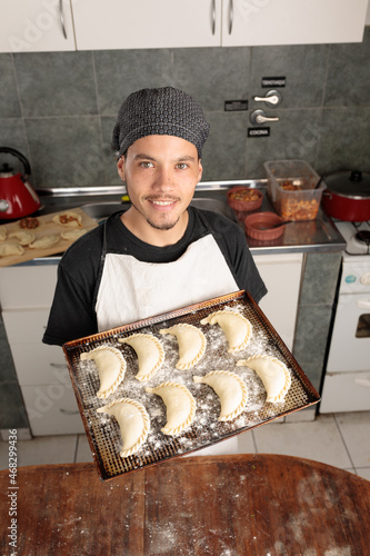 Young Latin American cook holds a baking tray with raw empanadas for cooking and looks at the camera smiling. Traditional South American food. Small entrepreneur works in his kitchen.