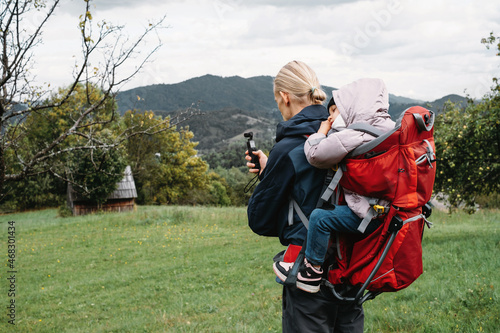 Father taking pictures while hiking with child in backpack. Vlogger man walking with sleepy kid in backpack in the cold mountains. Active time with tired girl outdoors 