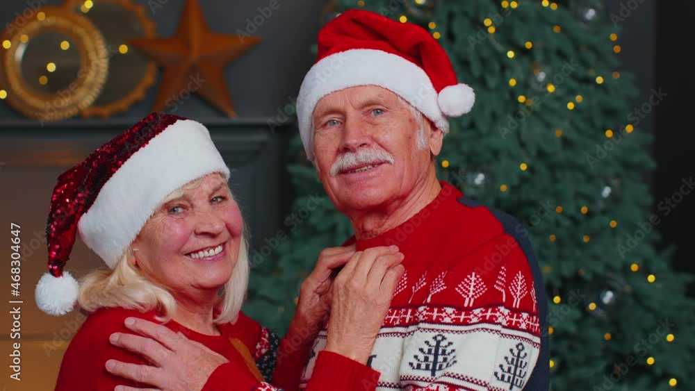 Portrait of happy senior couple in Santa Claus hats celebrating Christmas in cozy living room at home, looking into camera and hugging. Elderly people Grandparents celebrate Christmas. Holiday Mood Portrait of happy senior couple in Santa Claus hats celebrating Christmas in cozy living room at home, looking into camera and hugging. Elderly people Grandparents celebrate Christmas. Holiday Mood