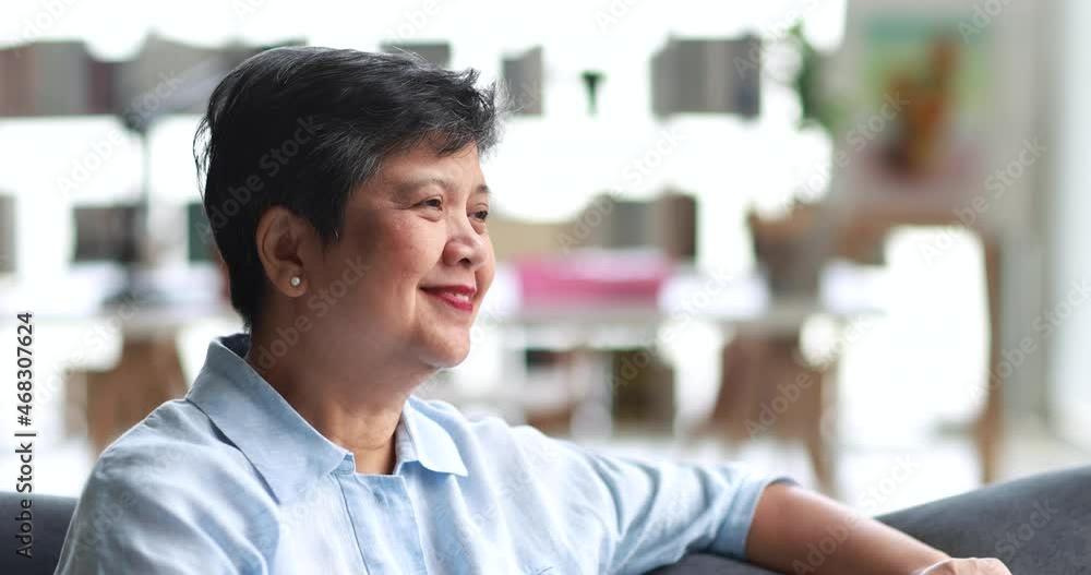 Positive mature Asian woman in blue shirt smiling and looking at camera while sitting on sofa in living room at home Positive mature Asian woman in blue shirt smiling and looking at camera while sitting on sofa in living room at home