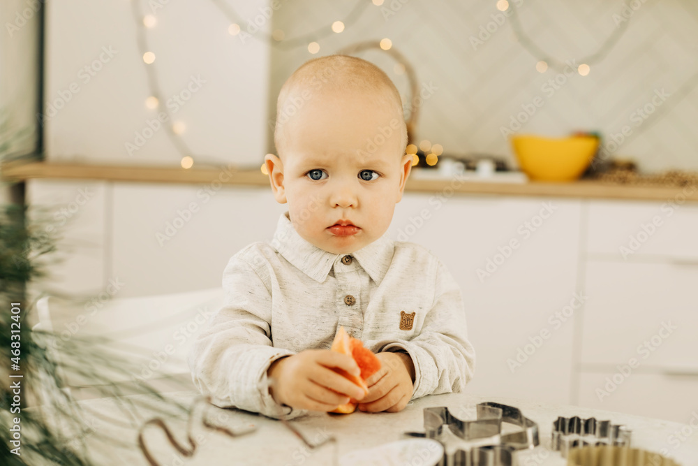 Naklejka premium A sad kid with a grapefruit in his hands is sitting at a table with cookie cutters on it. Preparing for the holidays, Christmas Eve or New Year's Eve
