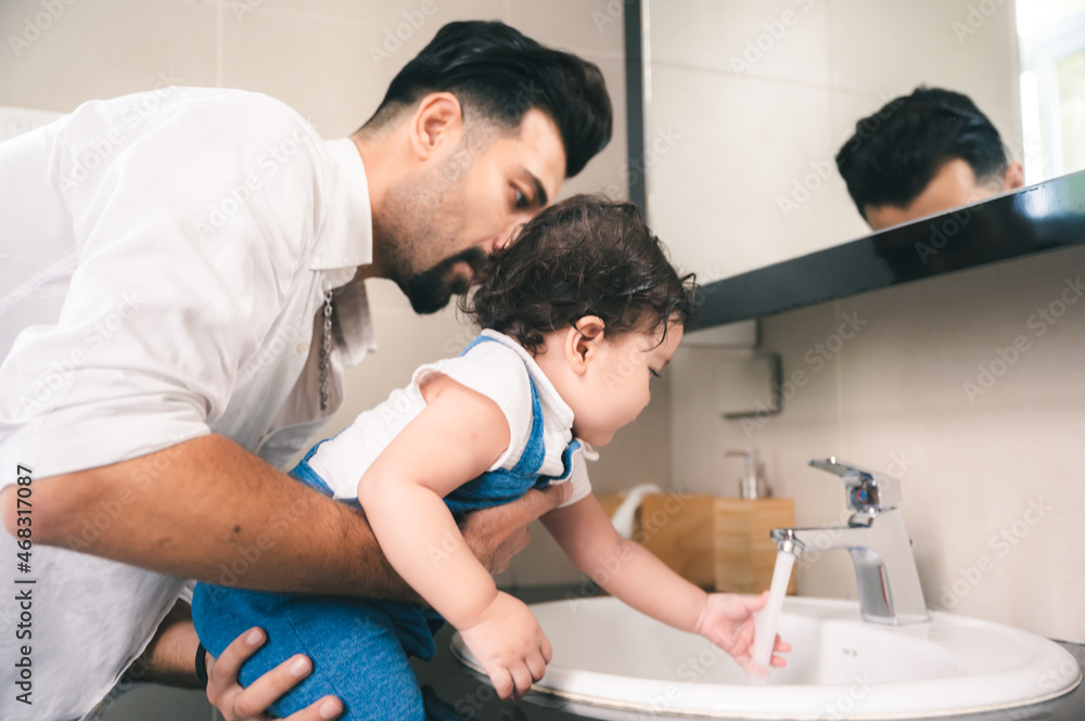 Handsome young father lifting small little daughter while training her about cleanliness by helping her wash her hands under running tap water in bathroom basin Handsome young father lifting small little daughter while training her about cleanliness by helping her wash her hands under running tap water in bathroom basin