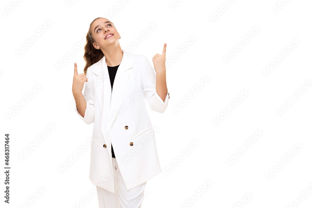 Photo of a beautiful cute adorable smiling brunette teenage girl with a ponytail in a stylish white jacket and white pants isolated on a white background shows a fingers up on a free space for text Photo of a beautiful cute adorable smiling brunette teenage girl with a ponytail in a stylish white jacket and white pants isolated on a white background shows a fingers up on a free space for text