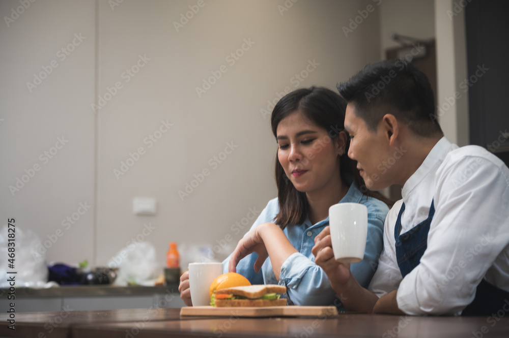 Cheerful and romantic young husband wearing apron drinking coffee with beautiful wife with sandwich and fruits at kitchen table while looking at each other Cheerful and romantic young husband wearing apron drinking coffee with beautiful wife with sandwich and fruits at kitchen table while looking at each other