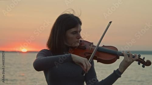 Young talented lady is playing violin skillfully, standing on rivershore and getting inspiration from pink sunrise, doing what she really loves, Foreground, Slow motion. photo