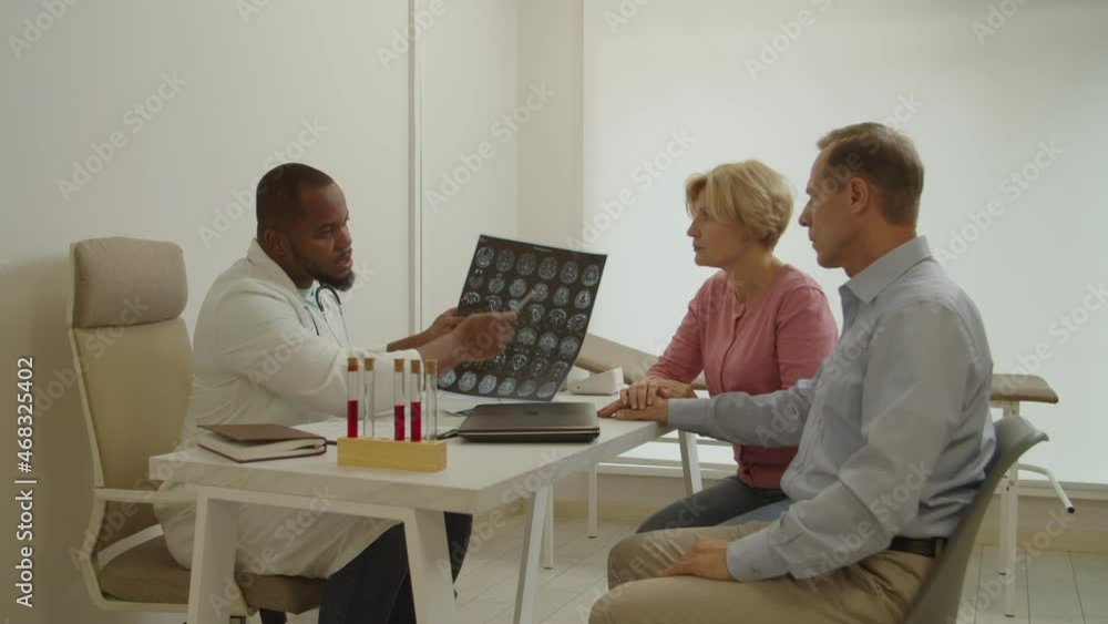 Professional black man doctor holding mri brain scan, explaining test results and treatment to worried middle aged male patient, supported by loving wife while mature couple visiting medical clinic. Professional black man doctor holding mri brain scan, explaining test results and treatment to worried middle aged male patient, supported by loving wife while mature couple visiting medical clinic.