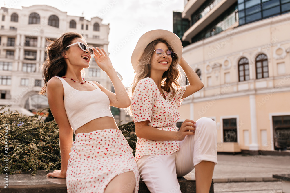 Two young beautiful smiling hipster girls posing on street background. In trendy summer clothes and hat positive models are having fun in sunglasses. Beauty, people emotions, summer vacation concept.
