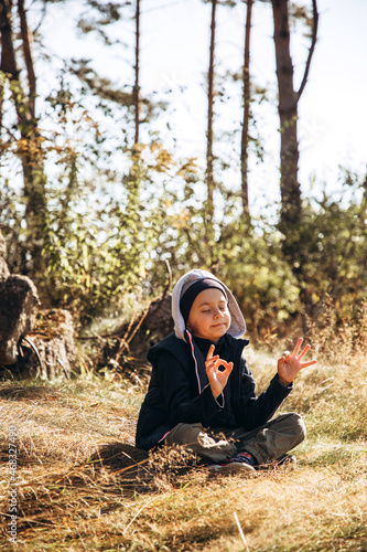 A boy in warm clothes meditates in the forest while sitting on the grass while walking. Portrait of a child