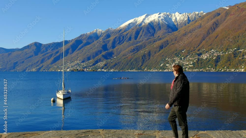 A Man Watching a Canoe on Alpine Lake Maggiore with Snow-capped Mountain with Clear Blue Sky in a Sunny Day in Ascona, Ticino, Switzerland. A Man Watching a Canoe on Alpine Lake Maggiore with Snow-capped Mountain with Clear Blue Sky in a Sunny Day in Ascona, Ticino, Switzerland.