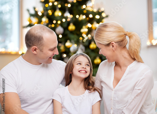 family, winter holidays and technology concept - portrait of happy mother, father and daughter sitting on sofa at home over christmas tree lights background © Syda Productions