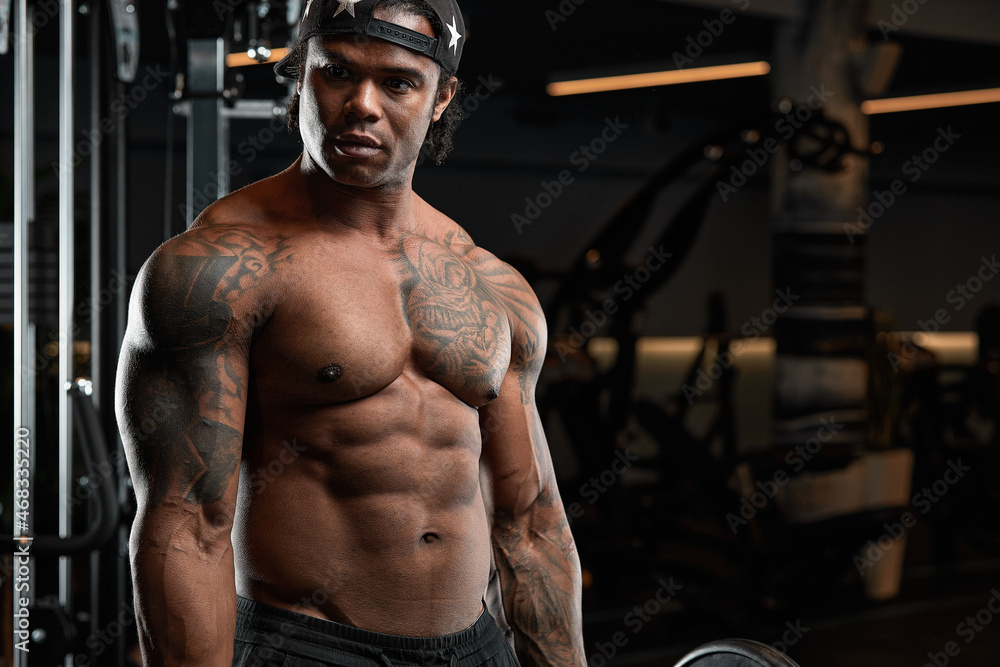 Strong and muscular dark skin man trains on modern equipment in gym. Portrait of muscular pumped up fitness trainer Strong and muscular dark skin man trains on modern equipment in gym. Portrait of muscular pumped up fitness trainer