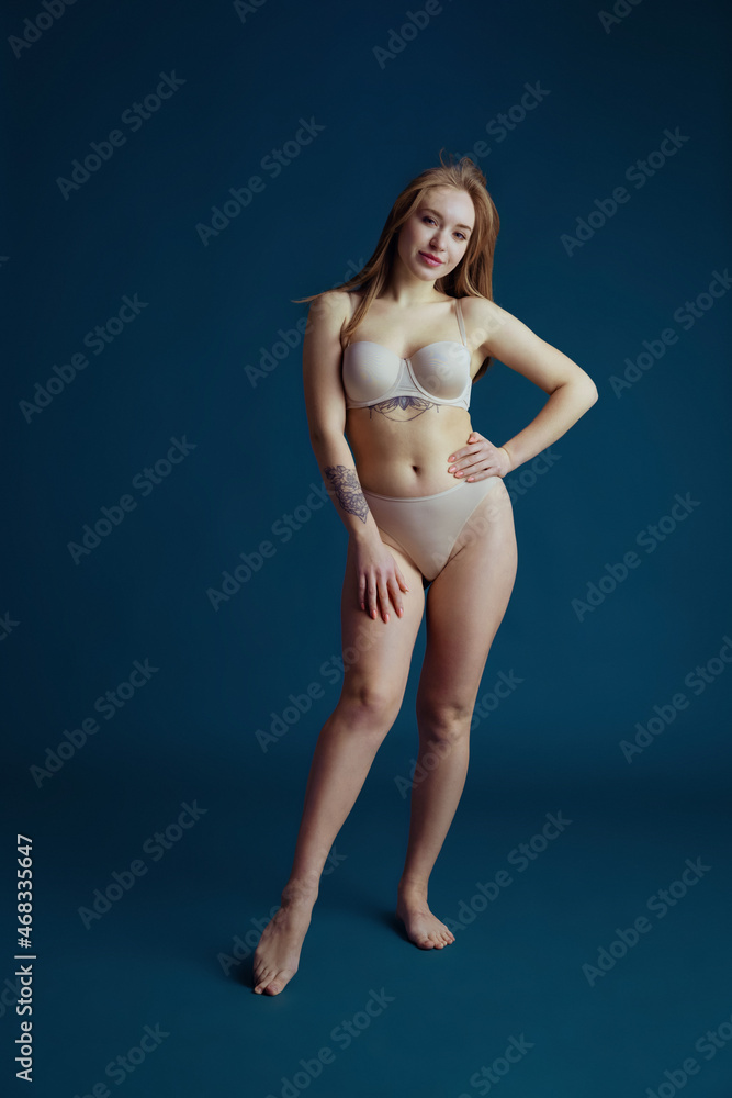 Portrait of young beautiful red-headed woman in inner wear posing isolated over blue studio background. Natural beauty concept.