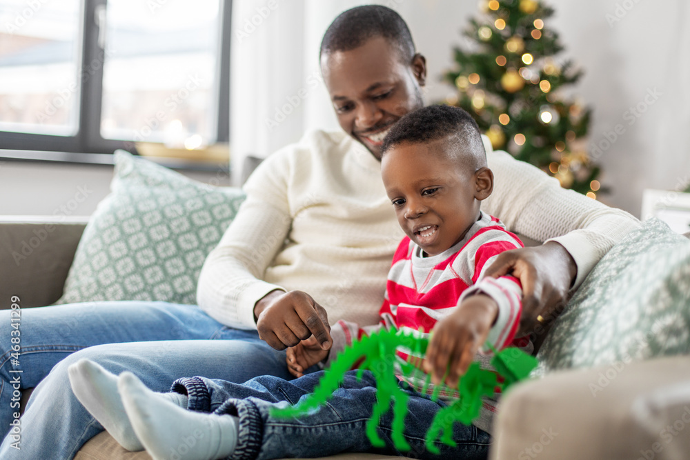 Fototapeta premium family, winter holidays and people concept - happy smiling african american father and baby son with dinosaur toy at home on christmas