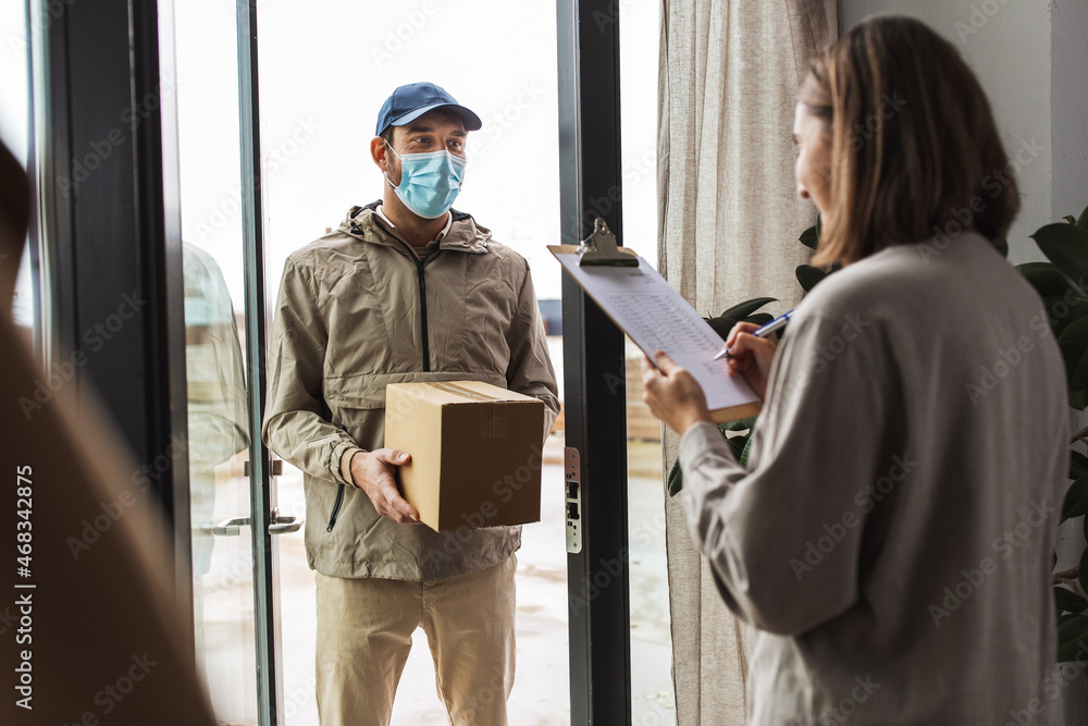 health protection, safety and pandemic concept - delivery man in face protective mask with parcel box and female customer signing papers at home health protection, safety and pandemic concept - delivery man in face protective mask with parcel box and female customer signing papers at home