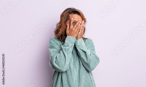 middle age woman feeling scared or embarrassed, peeking or spying with eyes half-covered with hands photo