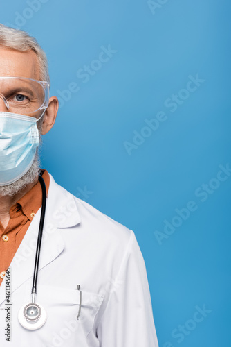 cropped view of middle aged doctor in white coat, medical mask and goggles on blue