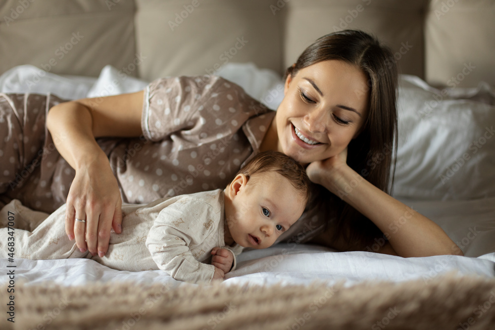 Happy mom plays with her newborn baby in the bedroom in beige colors on the bed.