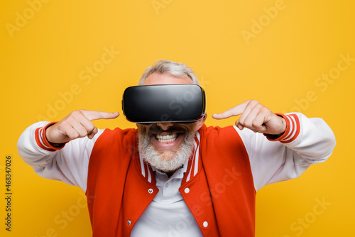 Foto cheerful middle aged man in bomber jacket pointing at vr headset isolated on yel