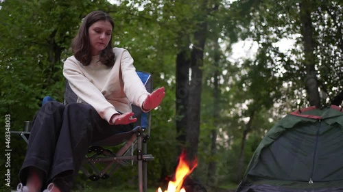 Lonely woman sits by the fire next to a tent in the woods. Rests and warms up on a hike. Twilight in nature