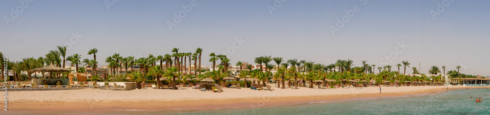 Hurghada, Egypt - September 22, 2021: Panoramic view of the sandy Egyptian beach with green palms. People relax, sunbathe on sun loungers and swim in the Red Sea.