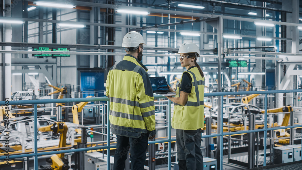Male Specialist and Female Car Factory Engineer in High Visibility Vests Using Laptop Computer. Automotive Industrial Manufacturing Facility Working on Vehicle Production. Diversity on Assembly Plant. Male Specialist and Female Car Factory Engineer in High Visibility Vests Using Laptop Computer. Automotive Industrial Manufacturing Facility Working on Vehicle Production. Diversity on Assembly Plant.
