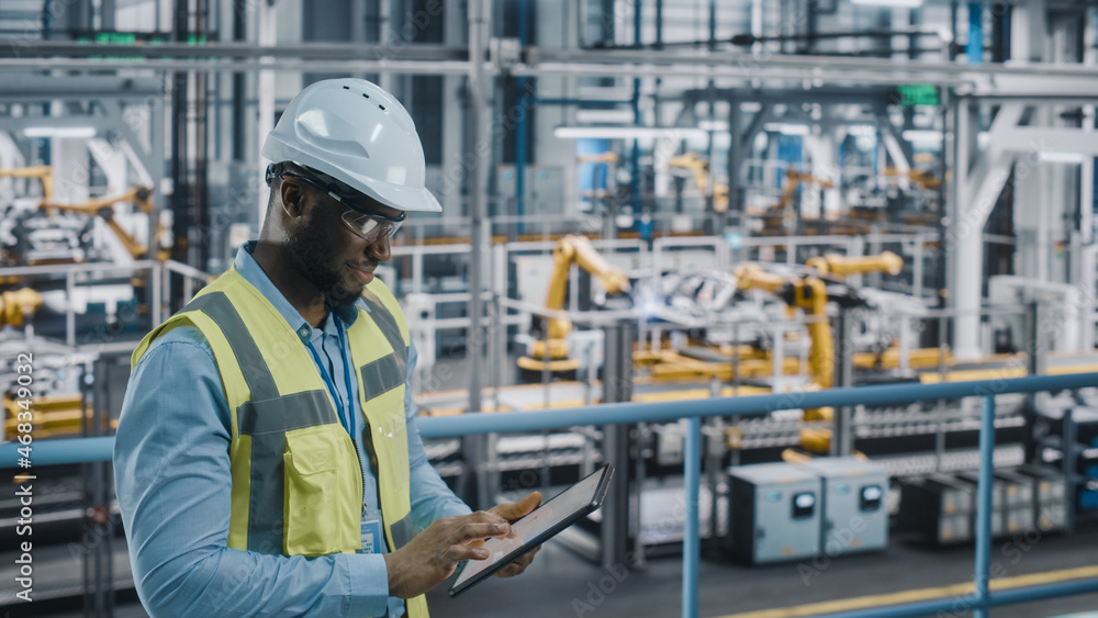 Happy African American Car Factory Engineer in High Visibility Vest Using Tablet Computer. Automotive Industrial Facility Working on Vehicle Production on Automated Technology Assembly Plant. Happy African American Car Factory Engineer in High Visibility Vest Using Tablet Computer. Automotive Industrial Facility Working on Vehicle Production on Automated Technology Assembly Plant.