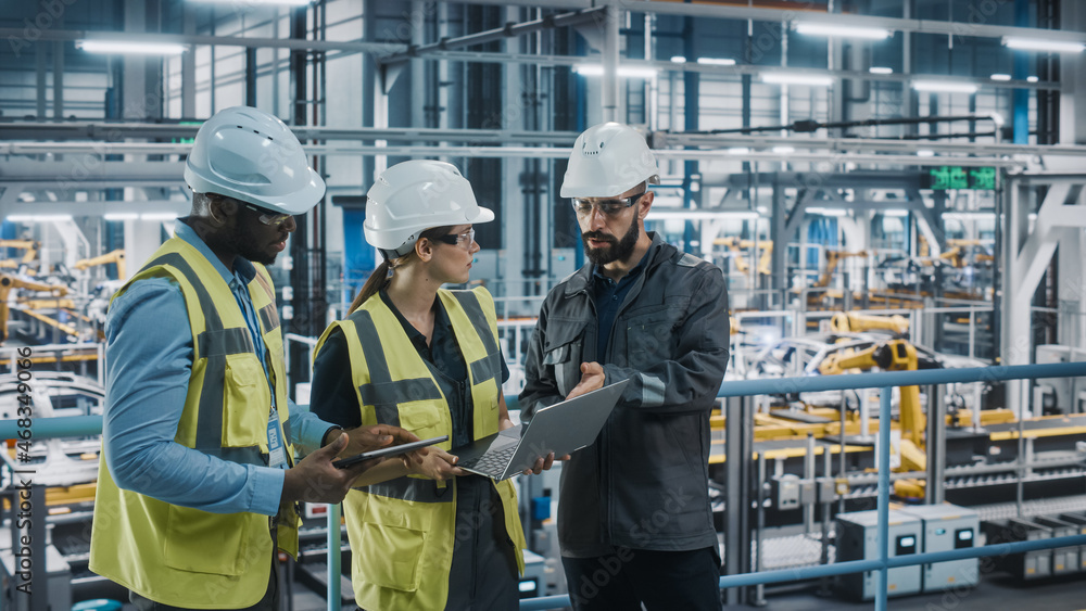 Young Diverse Team of Car Factory Specialists Working on Laptop and Tablet Computers. Engineers Discussing Automotive Industrial Manufacturing Technology on Modern Vehicle Assembly Plant. Young Diverse Team of Car Factory Specialists Working on Laptop and Tablet Computers. Engineers Discussing Automotive Industrial Manufacturing Technology on Modern Vehicle Assembly Plant.