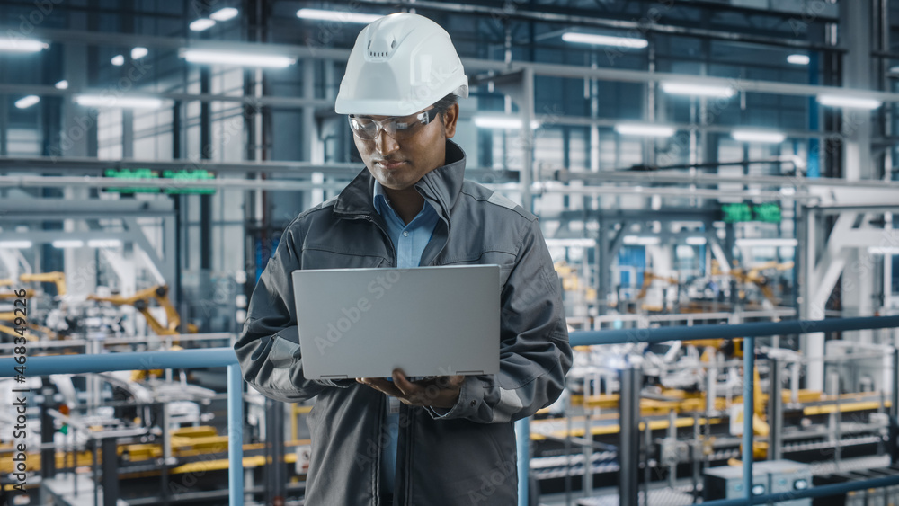 Portrait of Automotive Industry Engineer in Safety Uniform Using Laptop at Car Factory Facility. Happy Assembly Plant Multiethnic Man Specialist Working on Manufacturing Modern Electric Vehicles.