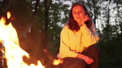 A young woman sits next to a fire in a camp. Warm and cozy campfire