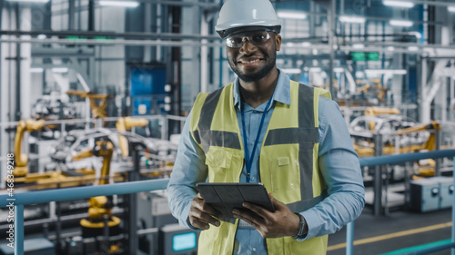 Portrait of Automotive Industry Engineer in Safety Uniform Using Tablet at Car Factory Facility. Happy Assembly Plant African American Man Specialist Working on Manufacturing Modern Electric Vehicles. © Gorodenkoff