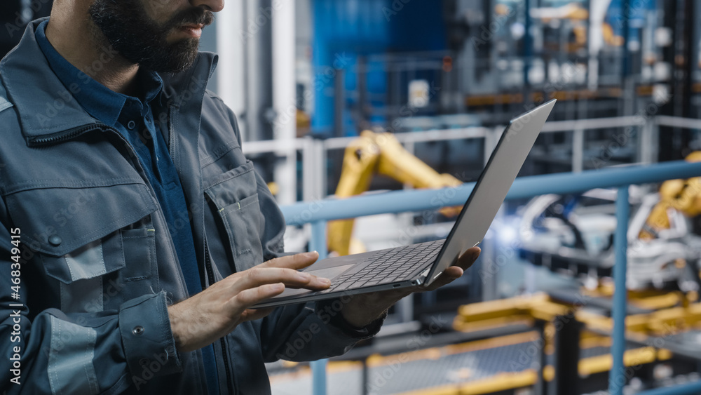 Close Up of Automotive Industry Engineer in Grey Uniform Using Laptop at Car Factory Facility. Professional Assembly Plant Specialist Working on Manufacturing Modern Electric Vehicles. Close Up of Automotive Industry Engineer in Grey Uniform Using Laptop at Car Factory Facility. Professional Assembly Plant Specialist Working on Manufacturing Modern Electric Vehicles.
