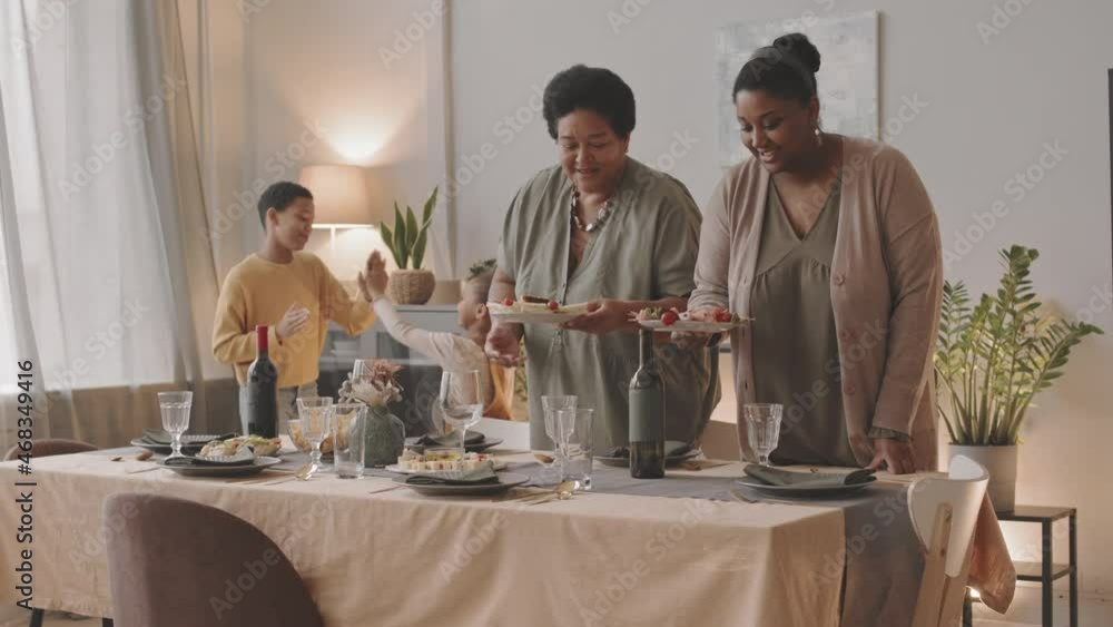 Titling up of two younger and older African American women putting plates with snacks on festive table and chatting in dining room at home, kids playing pat-a-cake on background Titling up of two younger and older African American women putting plates with snacks on festive table and chatting in dining room at home, kids playing pat-a-cake on background