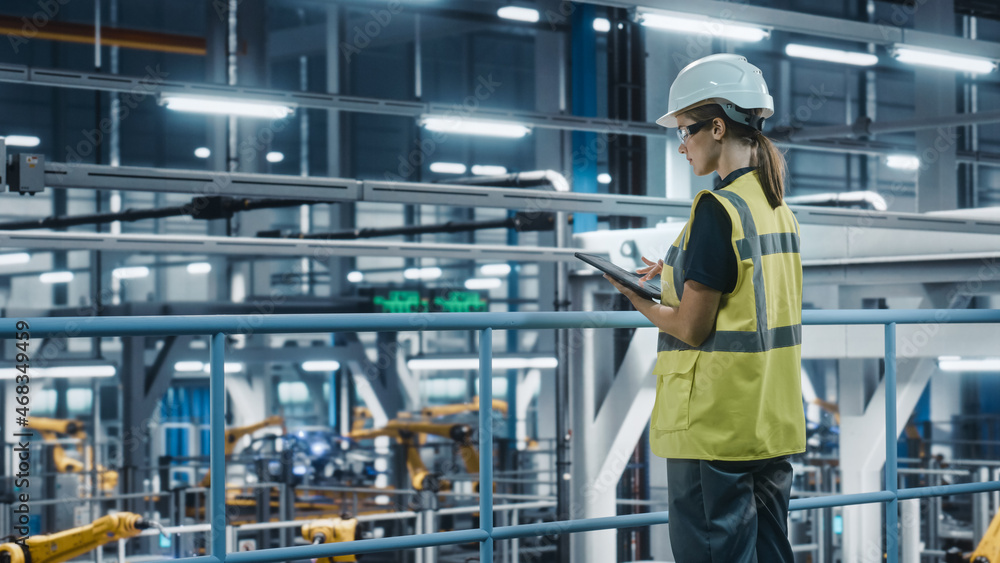 Female Car Factory Engineer in High Visibility Vest Using Tablet Computer. Automotive Industrial Manufacturing Facility Working on Vehicle Production with Robotic Technology. Automated Assembly Plant.