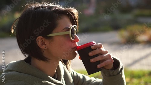 Close up portrait of young woman in casual sitting on the ground at the city park, drinking coffee from paper cup and enjoying the warm sunshine
