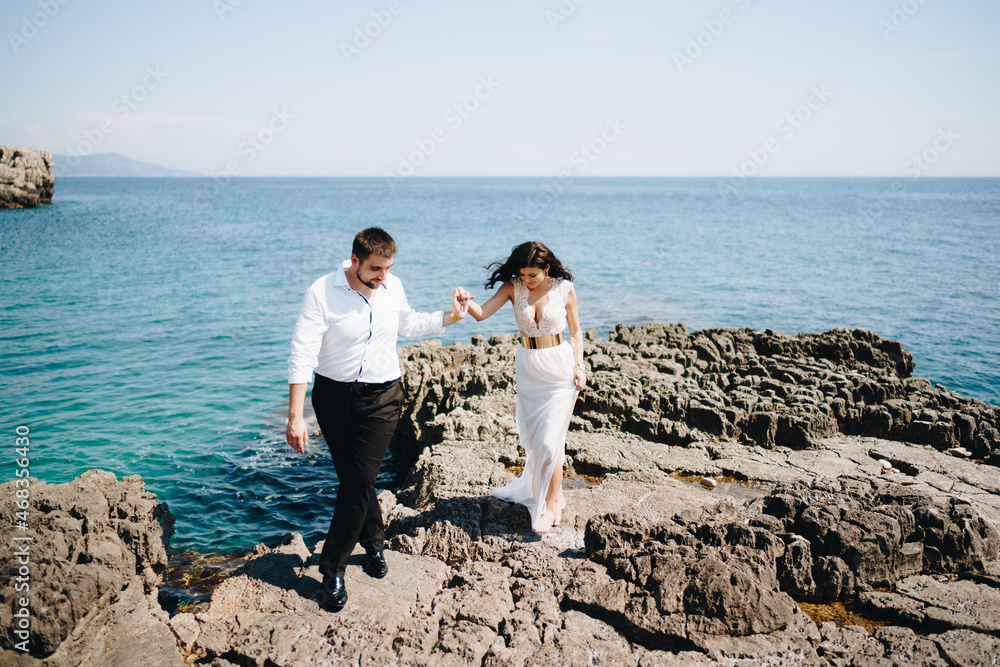 The bride and groom walk holding hands on the rocky shore 