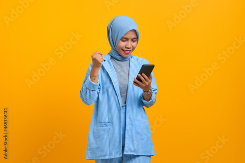 Cheerful young Asian woman holding mobile phone and celebrating success over yellow background