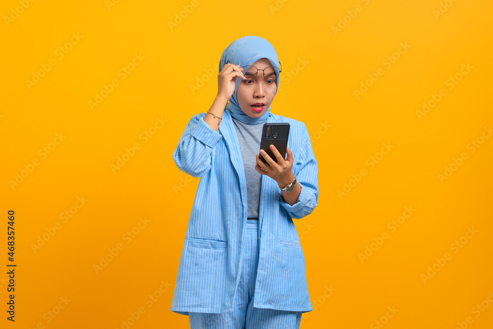Shocked young Asian woman using mobile phone and taking of glasses isolated over yellow background Shocked young Asian woman using mobile phone and taking of glasses isolated over yellow background