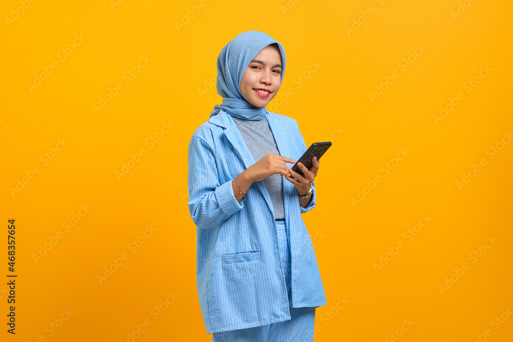 Smiling young Asian woman holding mobile phone and looking camera isolated over yellow background Smiling young Asian woman holding mobile phone and looking camera isolated over yellow background