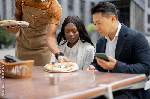 Multiracial business team having lunch and resting at outdoor cafe. Concept of teambuilding and corporate event. Waiter bringing pizza for clients. Businesspeople sitting at table with smartphones