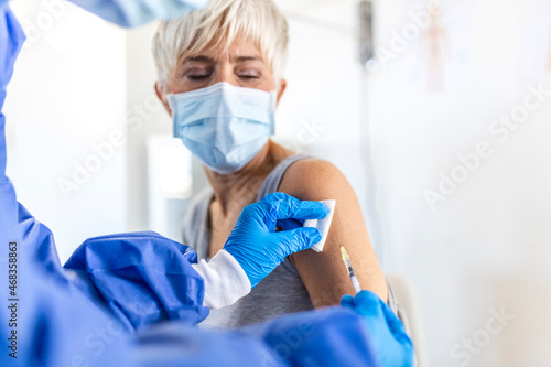 Close up of general practitioner hand holding vaccine injection while wearing face protective mask during covid-19 pandemic. Young woman nurse with surgical mask giving injection to senior woman.