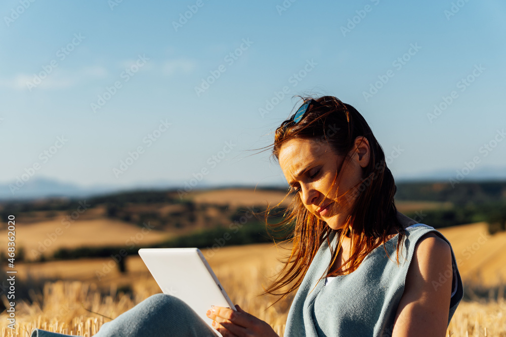 Middle-aged woman reading from tablet with sunglasses in her hair sitting in a cereal field. working. reading. Middle-aged woman reading from tablet with sunglasses in her hair sitting in a cereal field. working. reading.